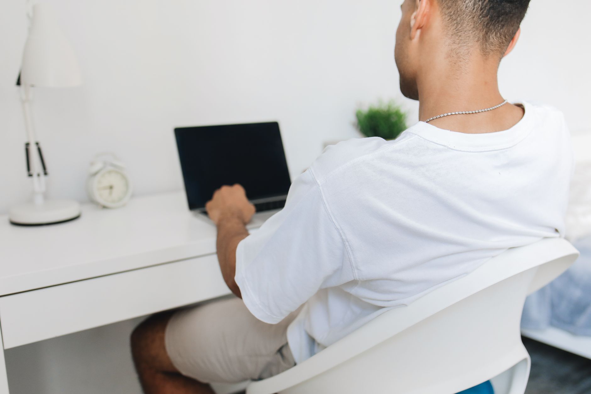 Une personne vue de dos, assise à un bureau blanc, tape sur un ordinateur portable.