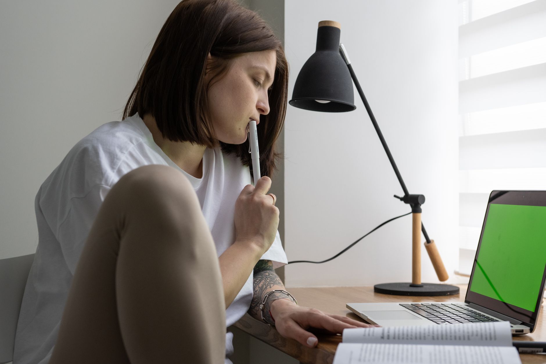 Une jeune femme pensive, un stylo près des lèvres, devant un ordinateur portable et un livre ouvert.
